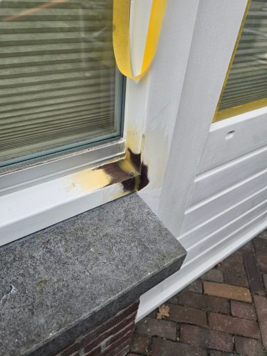 Beschadigde hoek van een raam met verkleuring en een gele lint eromheen.